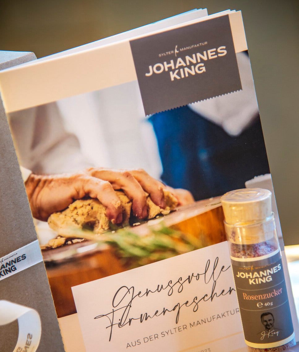 Eine Nahaufnahme eines Kochbuchs von Johannes King mit einem Bild von Händen, die auf dem Cover Essen zubereiten, und einer Flasche Rosenzucker an der Seite, auf deren Etikett der Name „Johannes King“ steht.