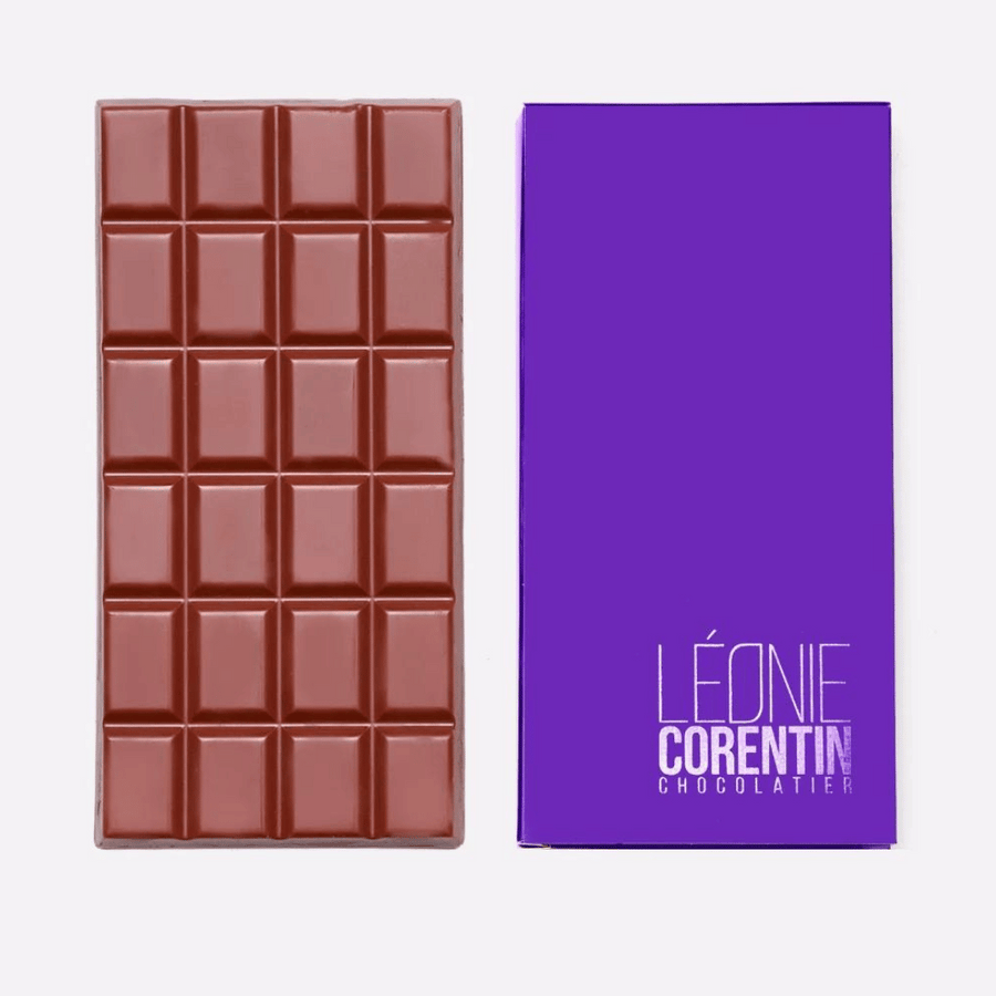 Eine Léonie Corentin Vollmilchschokolade 36 % mit Karamell mit segmentiertem Design wird neben ihrer violetten Verpackung platziert. Die Verpackung mit dem in Weiß aufgedruckten Text „LÉONIE COREN TIN CHOCOLATIER“ umschließt erstklassige Léonie Corentin für einen verwöhnenden Genuss.