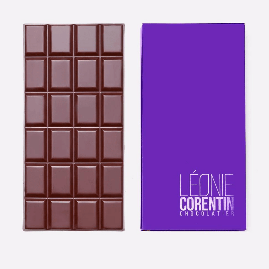 Neben einer violetten Schachtel ist eine Tafel Schokolade mit Gittermuster abgebildet. Auf der Schachtel steht in Weiß der Text „LÉONIE CORENTIN CHOCOLATIER“. Verwöhnen Sie sich mit dieser exquisiten Léonie Corentin Dunkle Schokolade 64 %, reichhaltiger dunkler Schokolade aus Madagaskar.