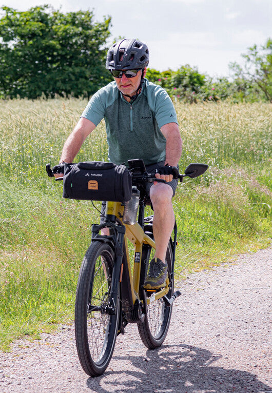 Ein Mann mit Helm fährt an einem sonnigen Tag mit hohem Gras auf beiden Seiten des Weges Fahrrad mit Vorderradtasche.
