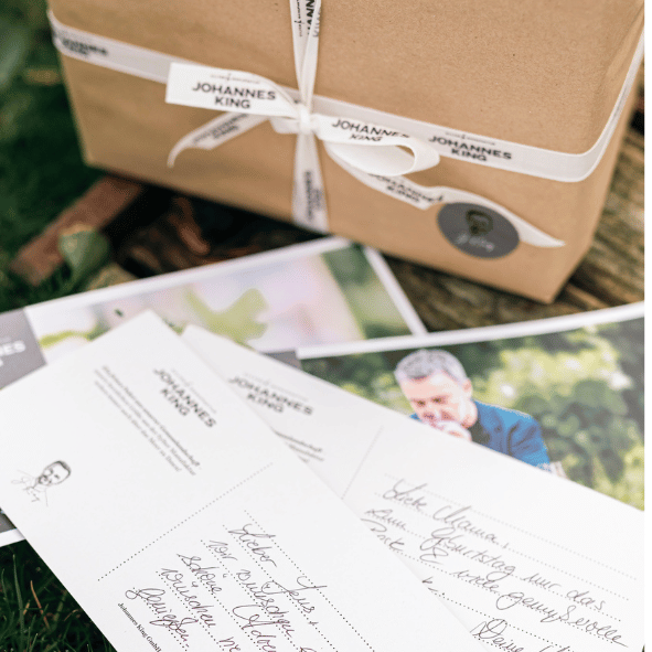Ein Stapel personalisierter Briefpapiere mit einem handgeschriebenen Brief auf der Oberseite liegt neben einem mit einer Schleife verschnürten Kartonpaket auf einer Grasfläche.