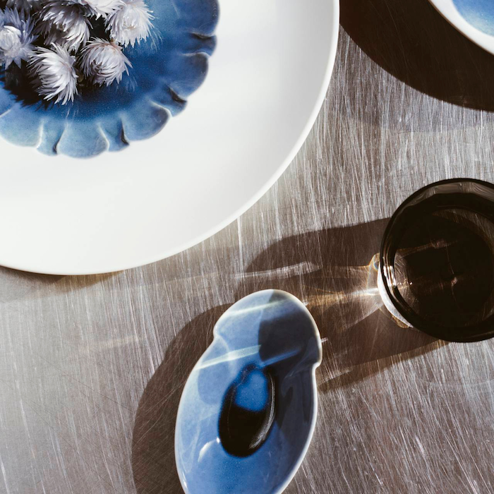 Ein Stilllebenfoto mit einer blauen Keramikkanne mit einer einzigartigen Glasur aus der Hering Berlin-Schale mit außenliegender freimodellierter Struktur Evolution – Blue Silent, einer Hering Berlin-Hohe Porzellanschale mit weißen Blumen und einem reflektierenden Glas.