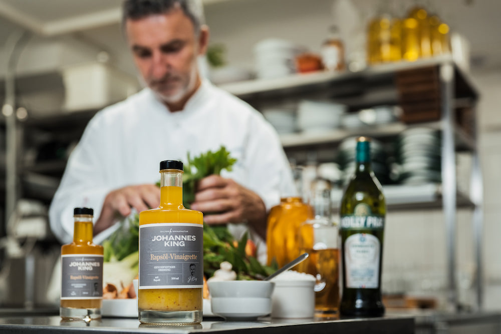 Ein Koch bereitet in einer professionellen Küche akribisch Speisen zu, im Vordergrund Flaschen mit Gourmetsaucen und Olivenöl.