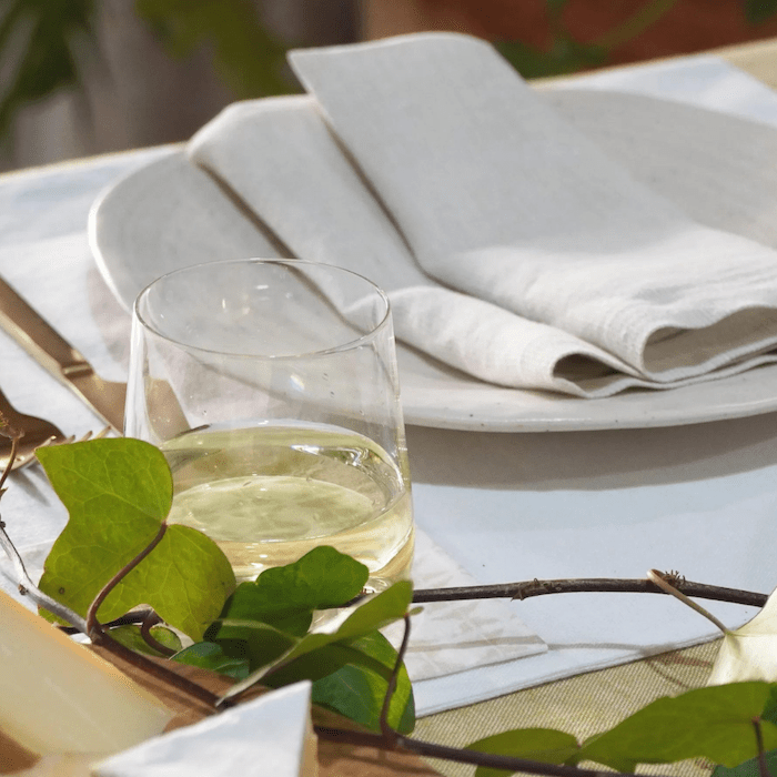 Ein Glas Weißwein auf einem mit einer Leinenserviette und elegantem Geschirr gedeckten Tisch, begleitet von einem Hauch Grün und ergänzt durch Dorothea Waydsch Kleine Brotzeit – Set.