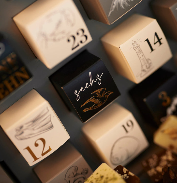 Eine Nahaufnahme eines Johannes King Schokoladen-Adventskalenders mit nummerierten Schubladen, wobei der Schwerpunkt auf der Schublade liegt, die mit der Nummer Sechs in eleganter Schrift und einer dekorativen Federillustration beschriftet ist.