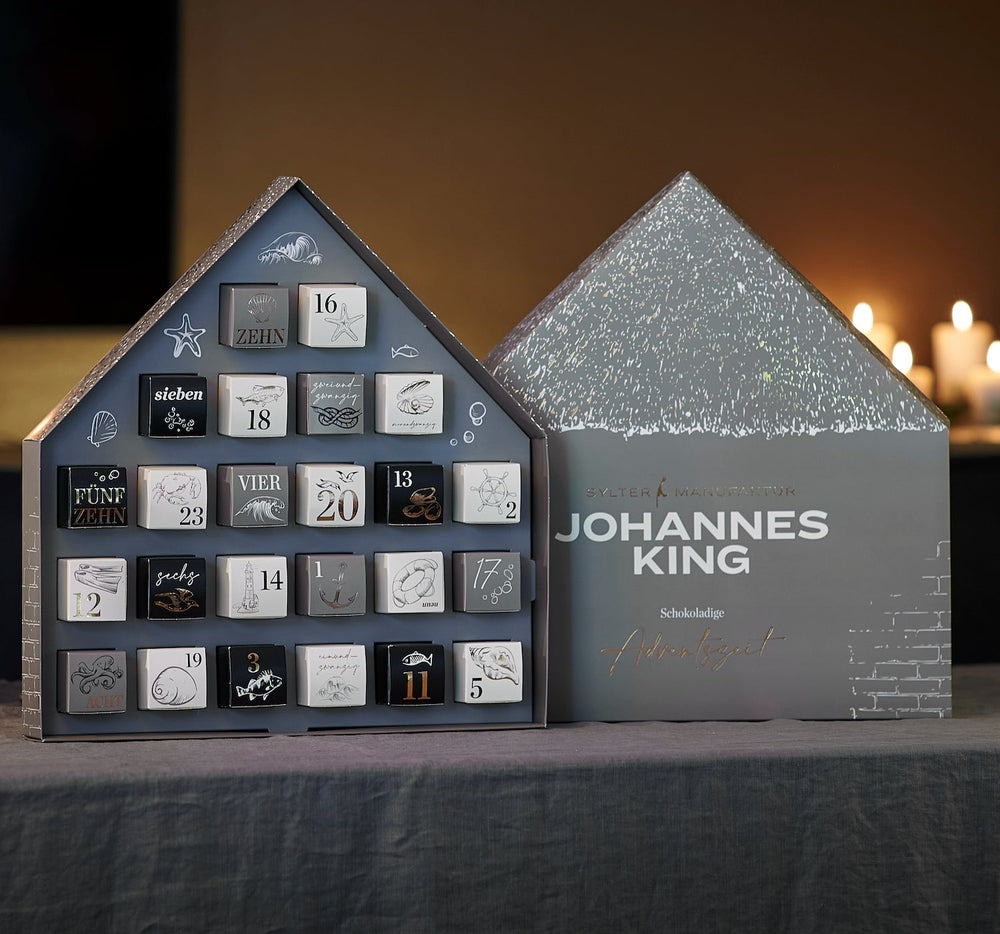Ein Adventskalender, der einem schneebedeckten Haus ähnelt, mit nummerierten Schubladen mit verschiedenen Artikeln von Kings Schokoladen und der Aufschrift „Johannes King“ auf der Vorderseite.