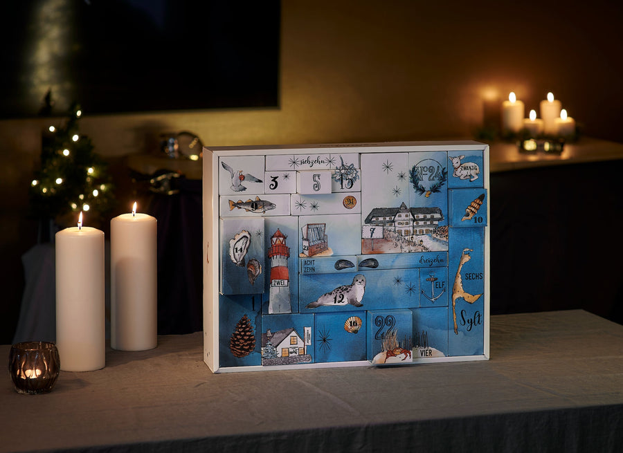 Ein Feinkost-Adventskalender 2023 *für Familien* von Sylter Manufaktur mit verschiedenen winterlichen und weihnachtlichen Illustrationen von Johannes King sitzt auf einem Tisch, mit ambientem Kerzen.