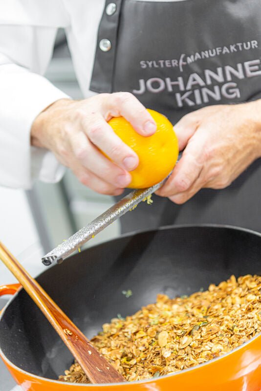 Eine Person mit einer schwarzen Schürze reibt die Schale einer Orange in einen Topf mit Sylter Manufaktur Granola Spezial mit Orangen, Zitronen & Ingwer (Stückliste).