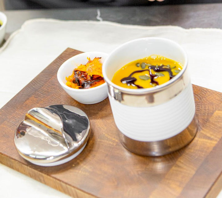 Eine Suppendose aus Porzellan mit Kürbissuppe, garniert mit einem Schuss dunklem Öl, präsentiert auf einem Holzbrett, dazu ein Schälchen mit Gewürzen und ein Suppenlöffel von der Sylter Manufaktur.