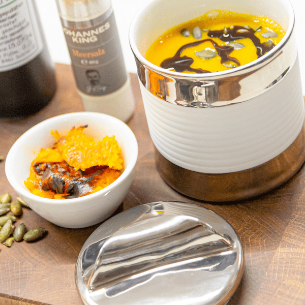 Eine Schüssel Kürbissuppe in einer Suppendose aus Porzellan der Sylter Manufaktur, garniert mit einem Schuss dunklem Öl, dazu ein Schälchen mit Kürbiskernen und Gewürzen, im Hintergrund eine Flasche.