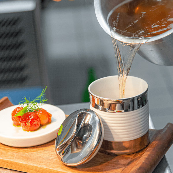 Eine Person gießt eine klare Brühe aus einer Metallkelle in eine Sylter Manufaktur Suppendose aus Porzellan neben einer garnierten Schüssel.
