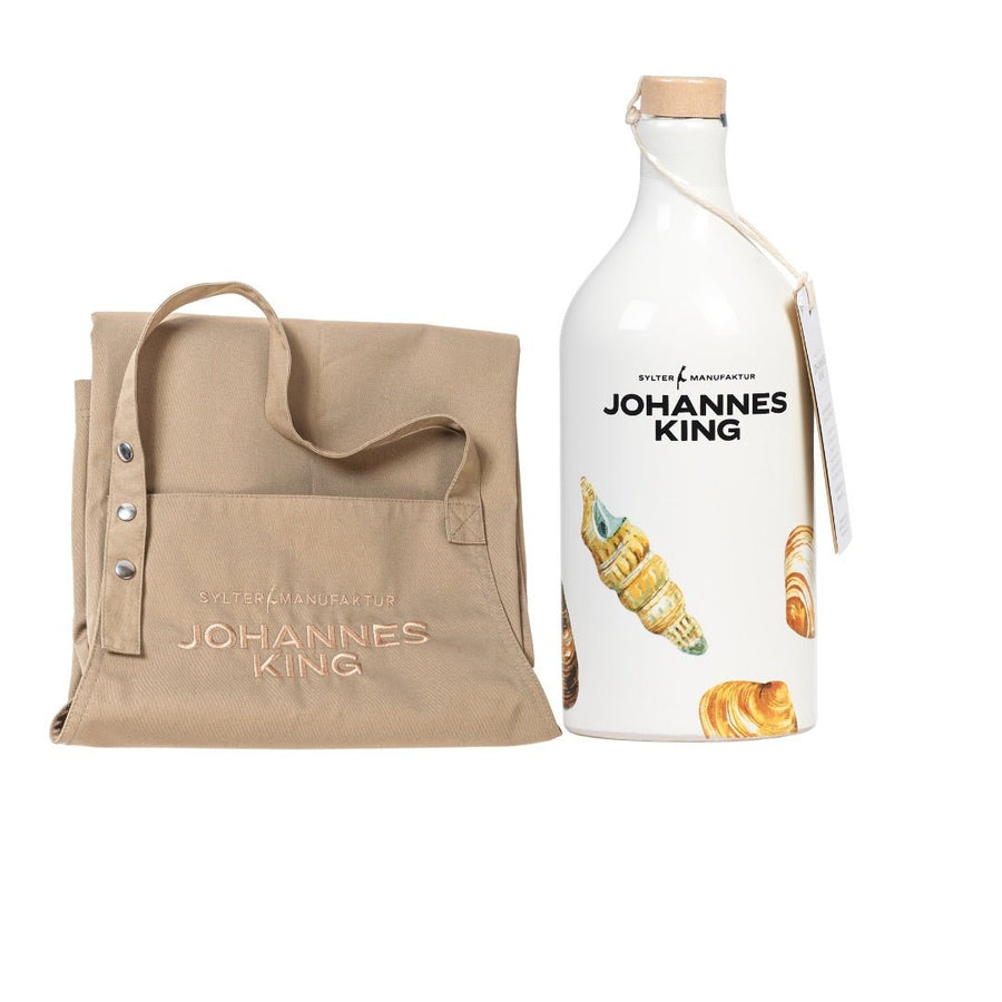Eine naturfarbene Tragetasche der Sylter Manufaktur mit „Johannes King“-Branding neben einer weißen Flasche Intensiv-Olivenöl derselben Marke, verziert mit Illustrationen von Croissants.