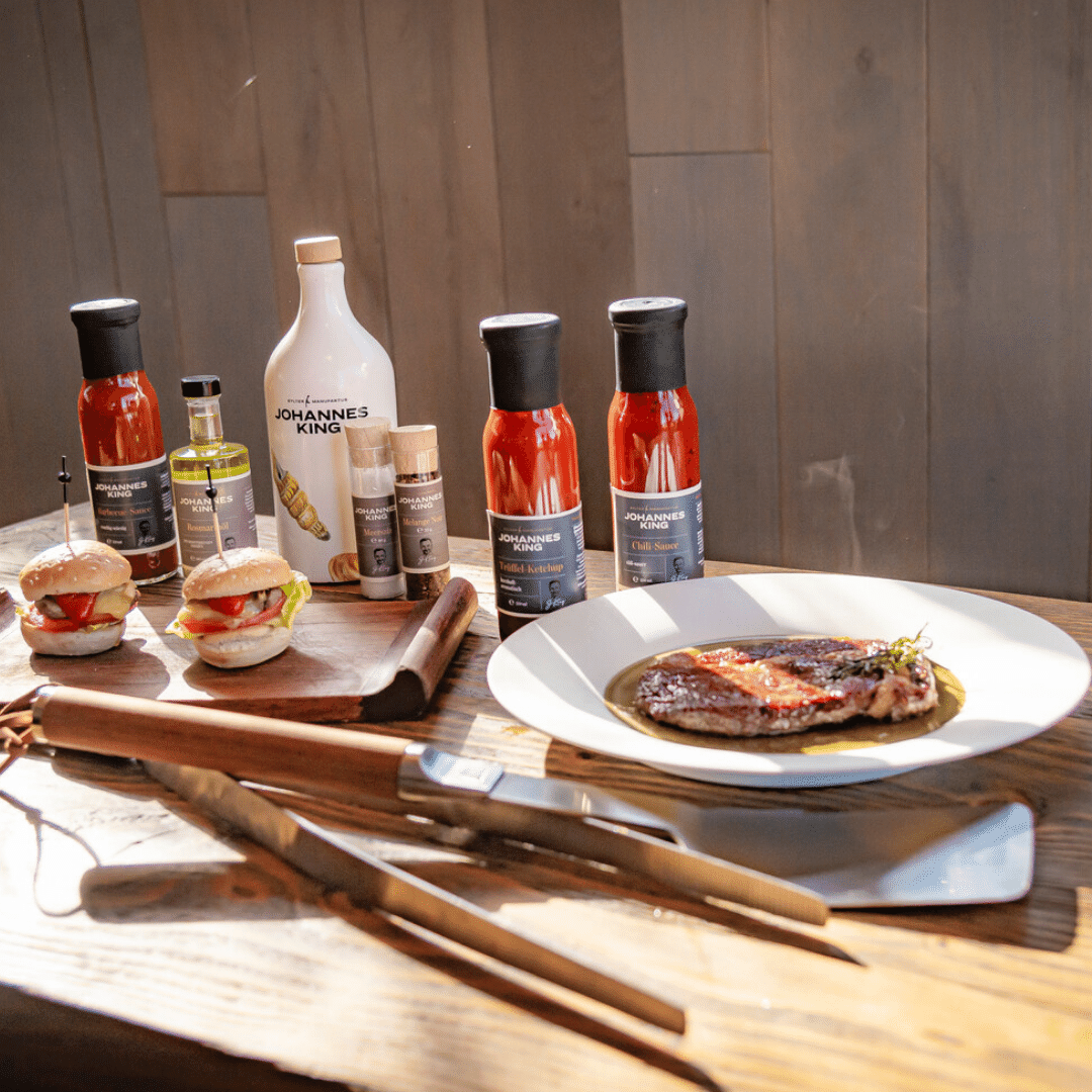 Verschiedene Grillsaucen, darunter das Würzige Grillsaucen-Set der Sylter Manufaktur, und Gewürze ordentlich auf einem Tisch angeordnet, daneben ein Teller mit einem gegrillten Steak und zwei Schiebern, auf dem Grillbesteck liegt.