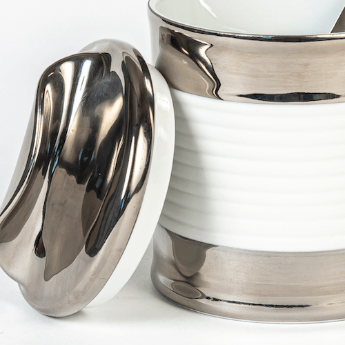 Ein polierter silberner Serviettenring, der an einem weißen Keramiktopf mit Sylter Manufaktur-Details lehnt.