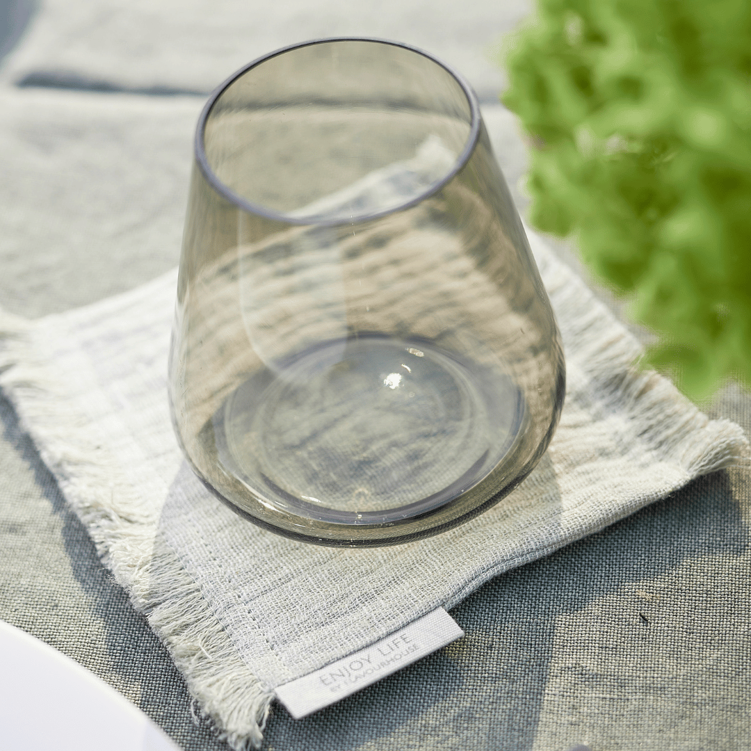 Ein leeres Glas liegt auf der Seite auf einer strukturierten Serviette mit dem Etikett „Genieße das Leben“, platziert auf einem Untersetzer-Set von Dorothea Waydsch mit einer grünen Pflanze.