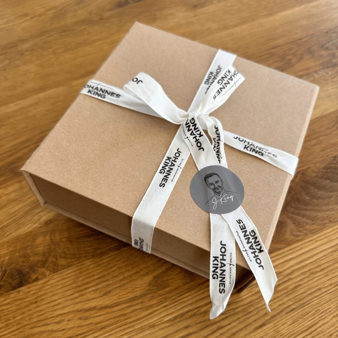 Ein verpackter Geschenkbox-Gutschein mit beigem Papier und einem weißen Band, bedruckt mit dem Text „Sylter Manufaktur Johannes King“ und einem runden Aufklebersiegel mit einem Silhouettenporträt.