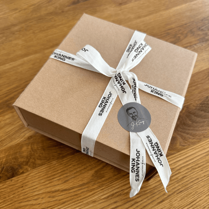 Ein verpackter Geschenkbox-Gutschein mit beigem Papier und einem weißen Band, bedruckt mit dem Text „Sylter Manufaktur Johannes King“ und einem runden Aufklebersiegel mit einem Silhouettenporträt.