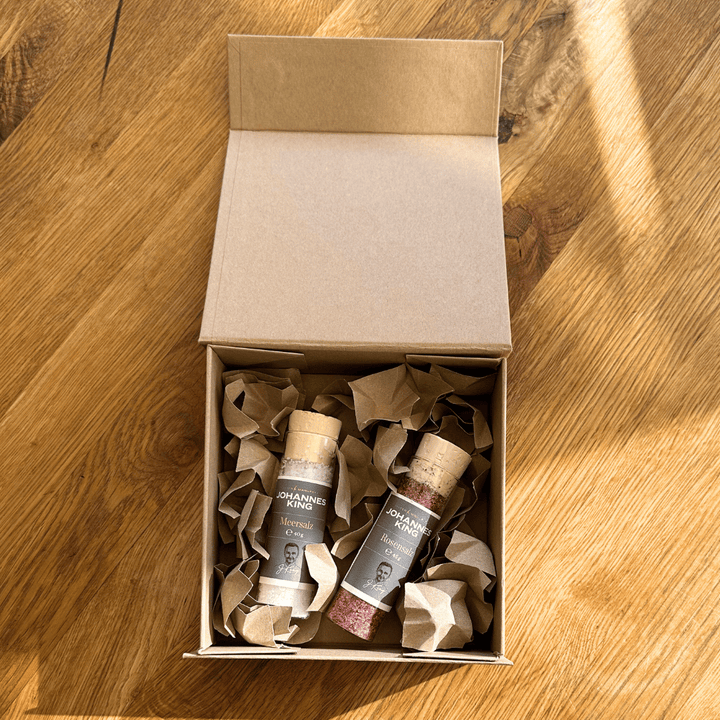 Ein offener Karton mit zwei Geschenkbox mit Salz-Duo-Flaschen der Sylter Manufaktur Johannes King, zum Schutz in zerknittertes Papier gewickelt, auf einem Holzboden im Sonnenlicht platziert.