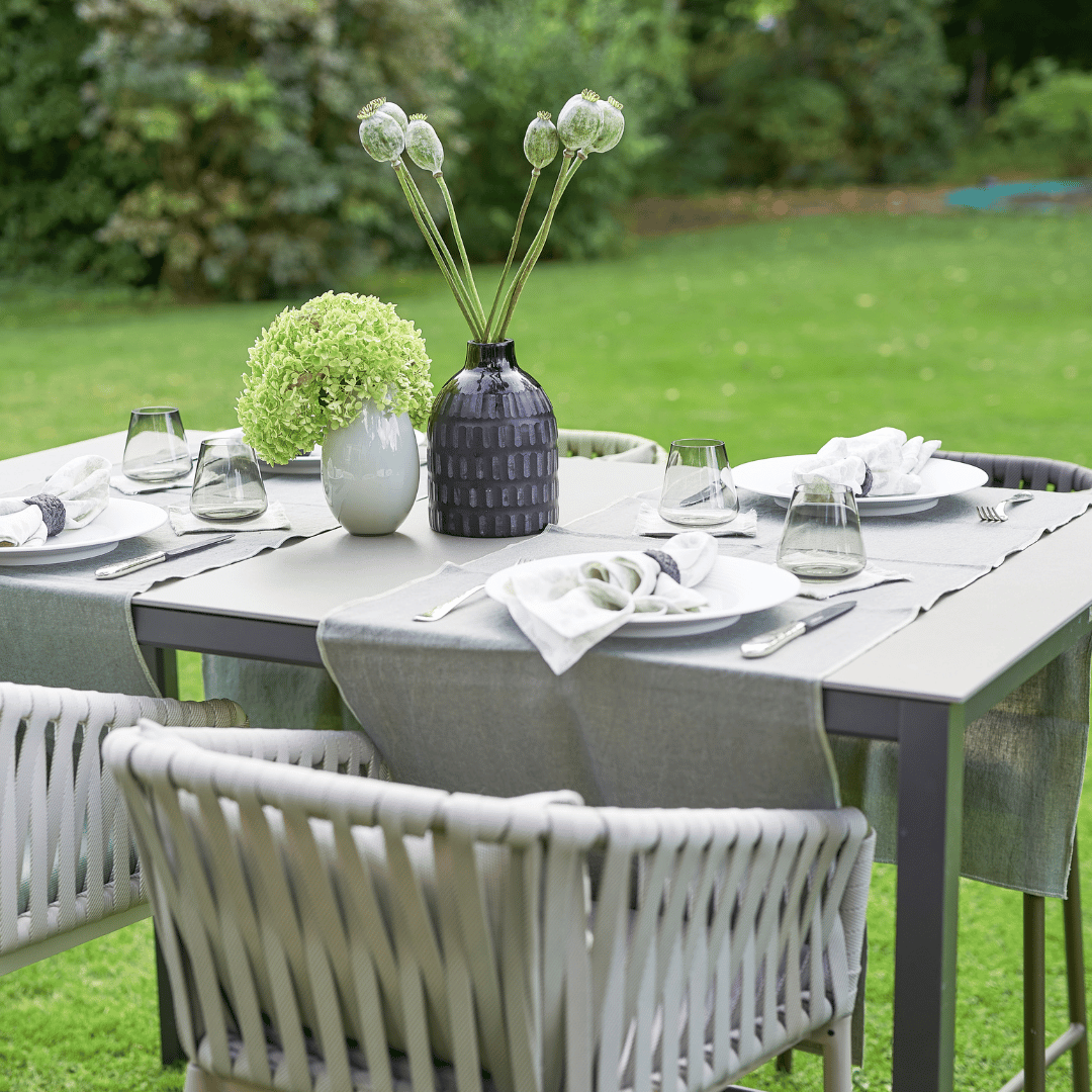 Ein Outdoor-Esstisch ist mit Tellern, Besteck, Gläsern und einem Tischläufer Dorothea Waydsch bestehend aus einem grünen Blumenarrangement in einem weißen V.