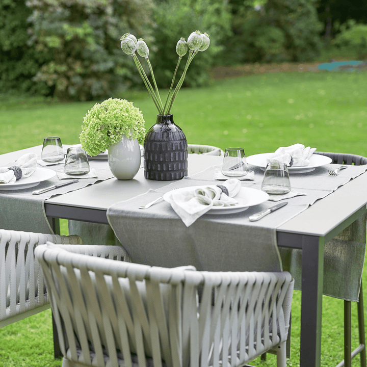 Ein Outdoor-Esstisch ist mit Tellern, Besteck, Gläsern und einem Tischläufer Dorothea Waydsch bestehend aus einem grünen Blumenarrangement in einem weißen V.