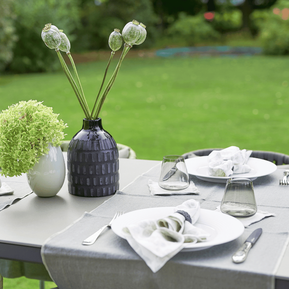 Ein Esstisch im Freien ist mit Tellern, Besteck und Gläsern gedeckt, mit einem grünen Blumenstrauß in einer weißen Vase und höheren Pflanzen in einer blauen Vase, dazu ein Tischläufer von Dorothea Waydsch.