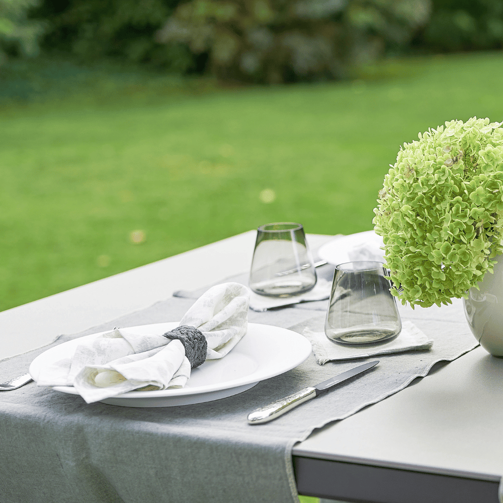 Ein Esstisch für zwei Personen im Freien mit weißen Tellern, Besteck, Weingläsern und einem grünen Hortensienaufsatz auf einer Tischdecke von Serviette Dorothea Waydsch mit einem Garten im Garten.