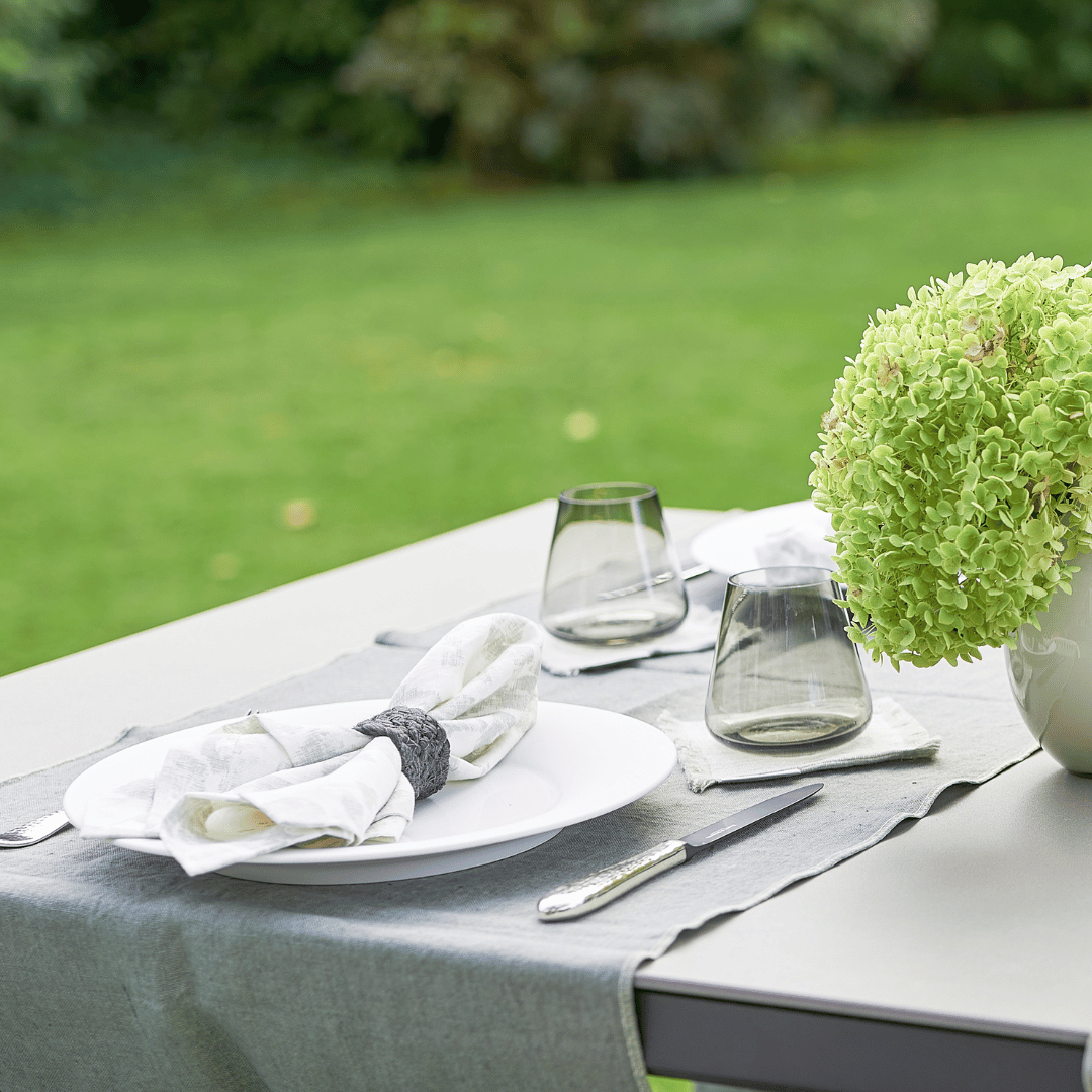 Ein Esstisch für zwei Personen im Freien mit weißen Tellern, Besteck, Weingläsern und einem grünen Hortensienaufsatz auf einer Tischdecke von Serviette Dorothea Waydsch mit einem Garten im Garten.