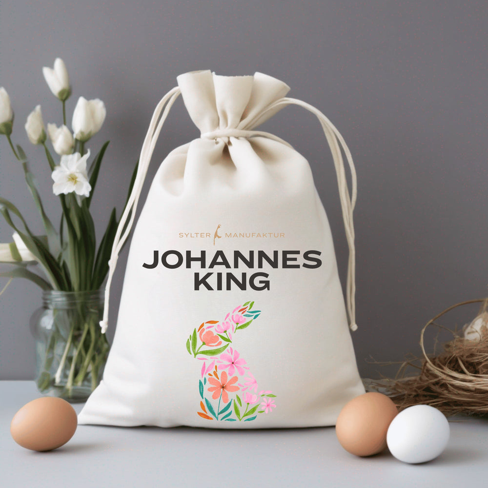 Ein personalisierter Osterbeutel mit Kordelzug und dem Aufdruck „Johannes King“ auf einem Tisch mit einem Strauß weißer Blumen auf der linken Seite, zwei Eiern im Vordergrund und einem Nest auf der rechten Seite vor grauem Hintergrund.
