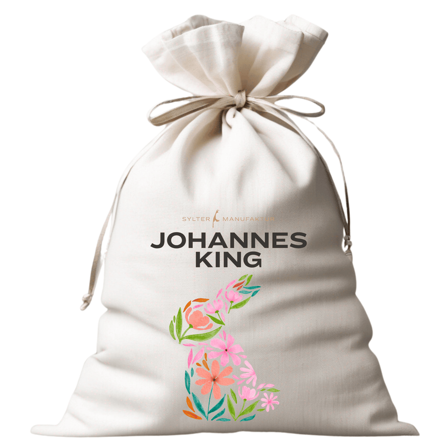 Ein weißer Canvas-Beutel mit Kordelzug, auf dem der Text „Sylter Manufaktur Johannes King“ über einem farbenfrohen Blumenmuster aufgedruckt ist und oben der zusätzliche Markentext „Osterbeutel“.