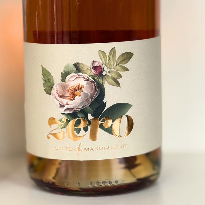 Eine Flasche mit einem floralen Etikett und einer elegant in Gold geschriebenen „Null“ hebt die Marke Sylter Manufaktur Johannes King hervor. Im Inneren offenbart die hell bernsteinfarbene Flüssigkeit subtile Nuancen von Kings Sylter Rosensecco *alkoholfrei* und bietet ein exquisites alkoholfreies Fruchtsecco-Erlebnis.