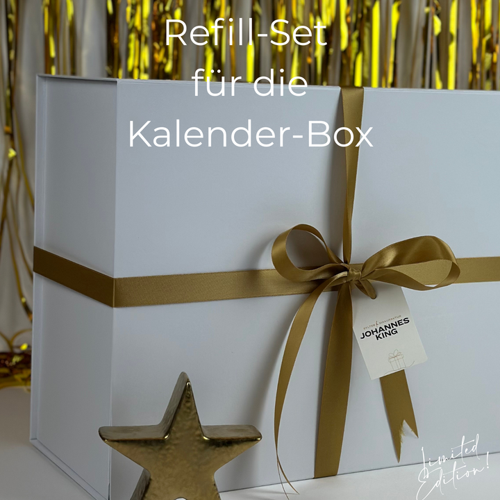 Eine weiße Schachtel mit goldenem Band und dem Schild "JOHANNES KING" steht vor goldenen Luftschlangen, davor ein goldener Stern. Überlagerter Text: "Nachfüll-Set* Adventskalender Box (2025), Sylter Manufaktur Johannes King.