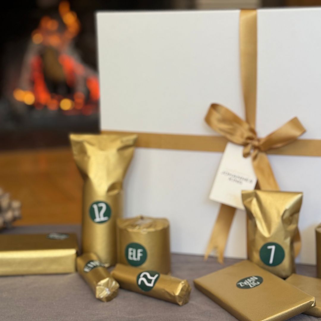 Auf einem Tisch steht ein Arrangement des Adventskalenders 2024 der Sylter Manufaktur Johannes King mit goldverpackten Geschenken mit Zahlen und Buchstaben. Eine große weiße Schachtel mit einer goldenen Schleife begleitet die Kollektion, während ein gemütliches Feuer im Hintergrund die warme Atmosphäre verstärkt.