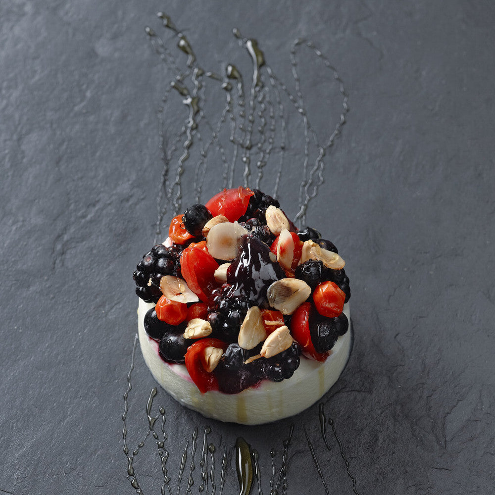 Ein cremiges Dessert, garniert mit gemischten Beeren und Nüssen, beträufelt mit dunklem Sirup auf dunklem Schiefergrund.