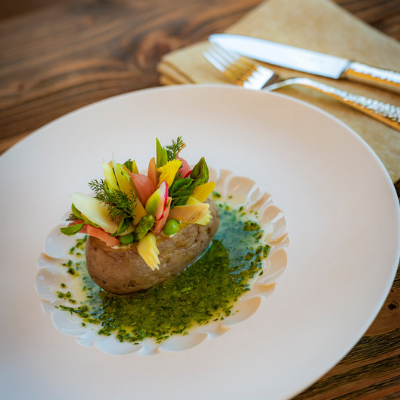 Eine Ofenkartoffel, garniert mit verschiedenen bunten Gemüsebeilagen, liegt auf einem weißen Teller mit Basilikum-Öl-Dressing. Daneben liegen eine gefaltete Serviette und Besteck.