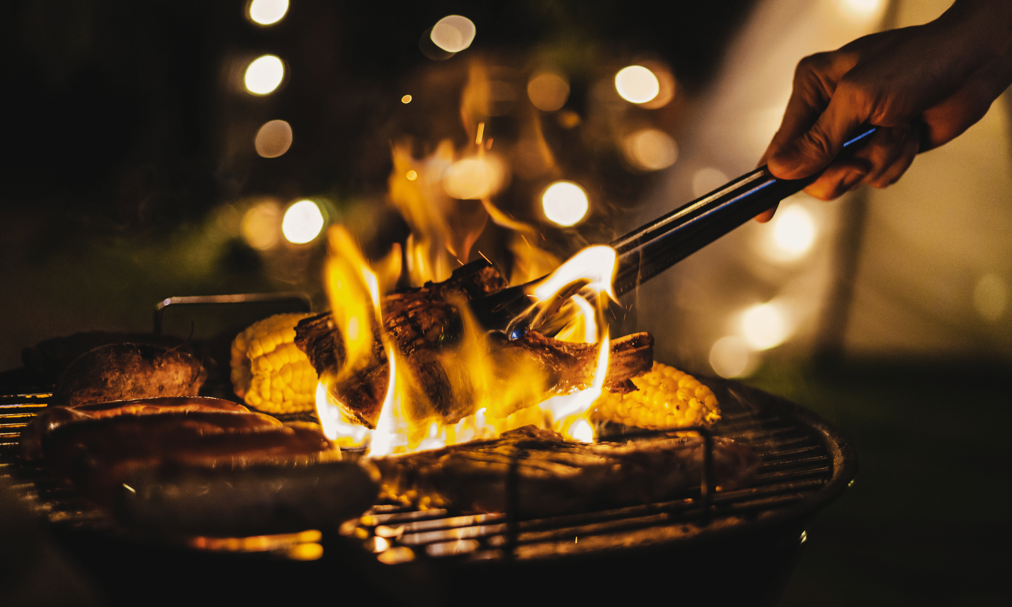 Eine Person grillt nachts verschiedene Lebensmittel auf einem Grill mit sichtbaren Flammen und schafft so eine warme und einladende Atmosphäre.