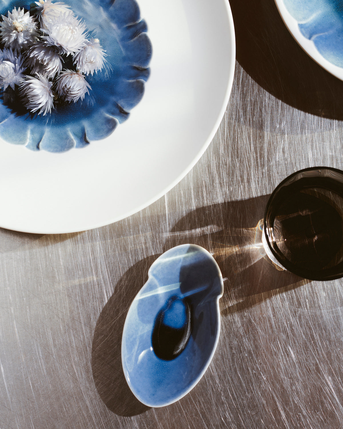 Eine blaue Keramikschale mit Blumenmotiv und weißen und blauen Blumen steht neben einem ovalen Keramikstück mit glänzender Oberfläche und einer dunklen Flüssigkeit in der Mitte, auf einer strukturierten Oberfläche, in deren Rahmen teilweise ein Metallbecher sichtbar ist.