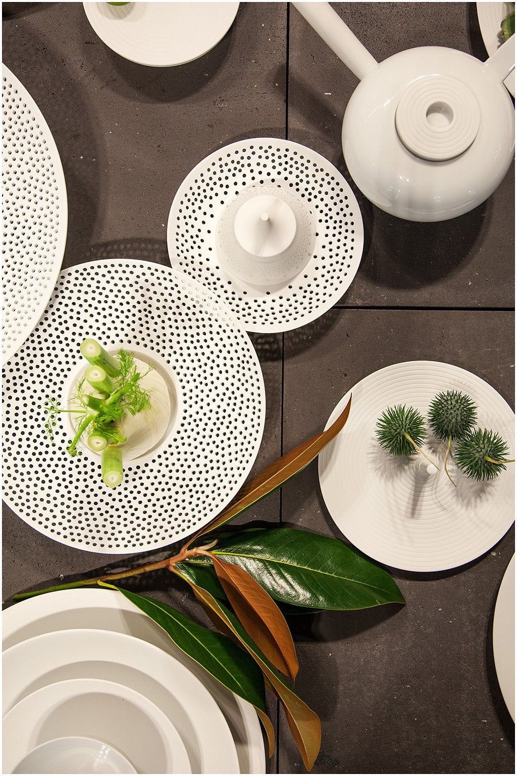 Eine stilvolle Tischdekoration mit einer Sammlung von weißem Geschirr mit schwarzen Tupfenmustern, ergänzt durch grüne Dekopflanzen und eine weiße Teekanne.