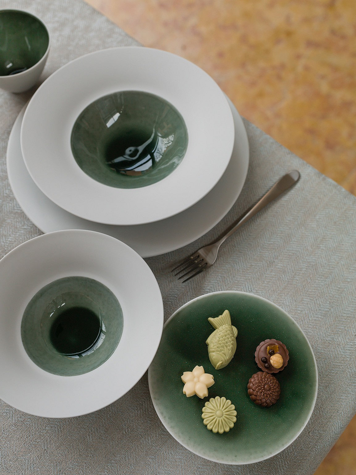 Ein Set Keramikgeschirr auf einem Tisch, bestehend aus zwei Tellern und zwei Schalen mit grüner Glasur, dazu eine Gabel und vier kleine Deko-Süßigkeiten auf einem Teller.