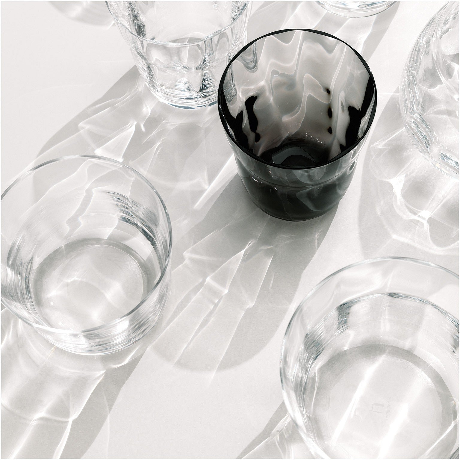 Mehrere transparente Glasgefäße sind auf einer lichtreflektierenden Oberfläche angeordnet, wobei das Lichtspiel komplizierte Schatten wirft, und eine dunkel getönte Glasschale sticht unter den klaren hervor.