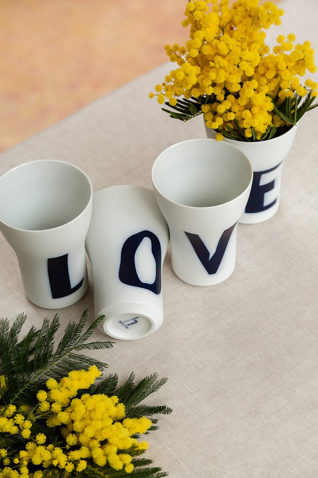 Vier weiße zylindrische Vasen mit Buchstaben darauf, die „Liebe“ bedeuten, wobei eine Vase umgeworfen wurde und in zwei der aufrecht stehenden Vasen gelbe Blumen arrangiert waren.