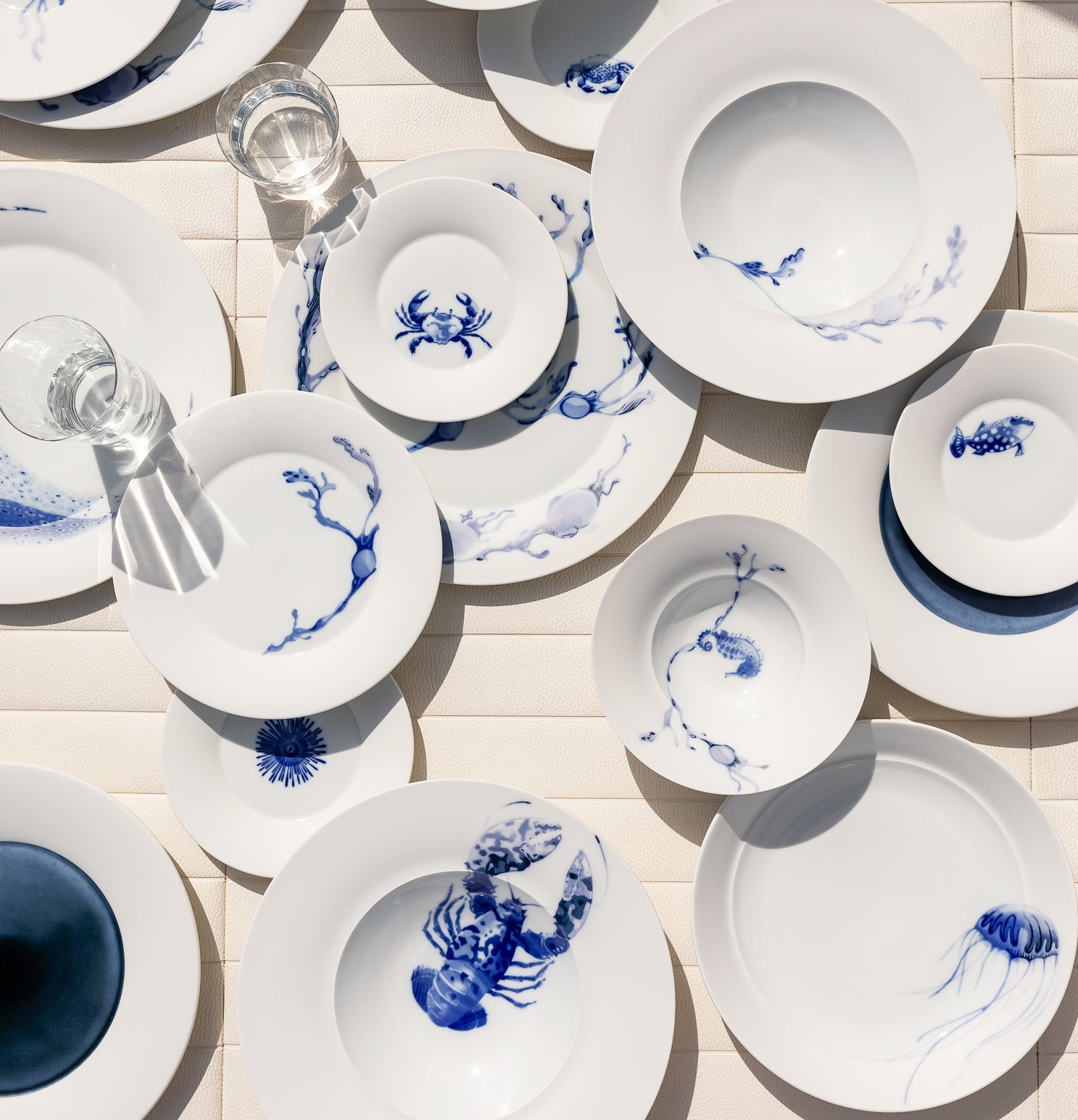 Weißes Geschirr mit blauen Mustern auf einem Tisch mit weißer Tischdecke und Sonnenlicht, das Schatten wirft.