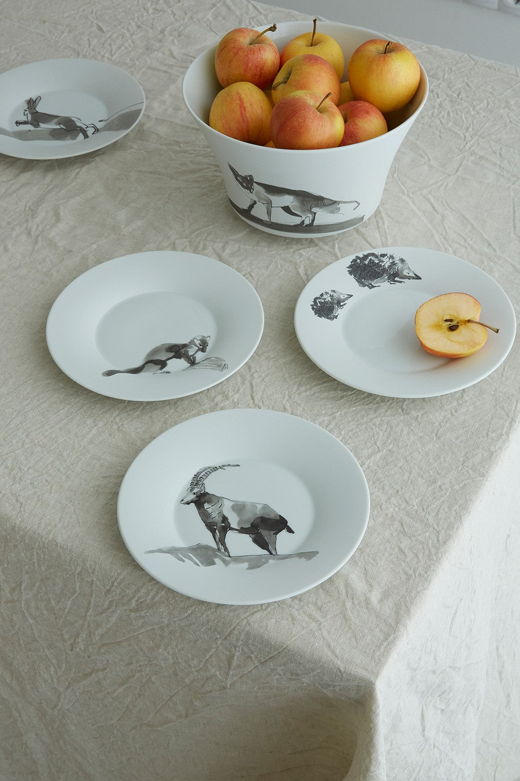 Ein Set Keramikgeschirr mit Tierillustrationen auf einem Tisch mit einer Tischdecke in neutralen Farben. Auf einem der Teller steht eine Schüssel mit Äpfeln und ein geschnittener Apfel.