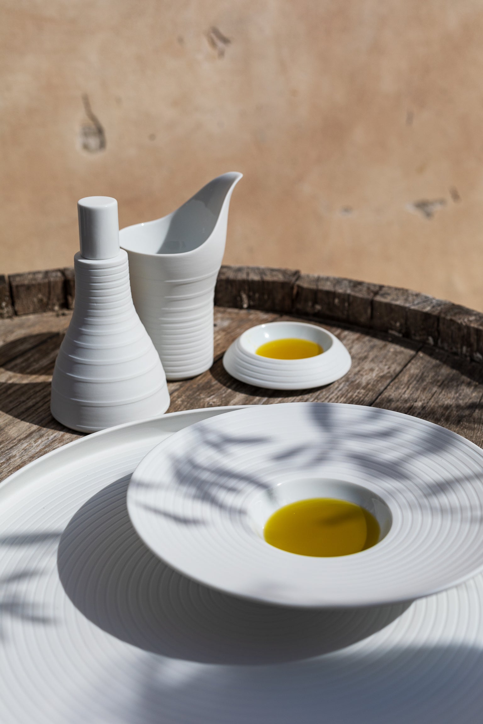 Weißes Keramikgeschirr mit Olivenöl auf einer Holzoberfläche platziert, das im natürlichen Licht Schatten wirft.