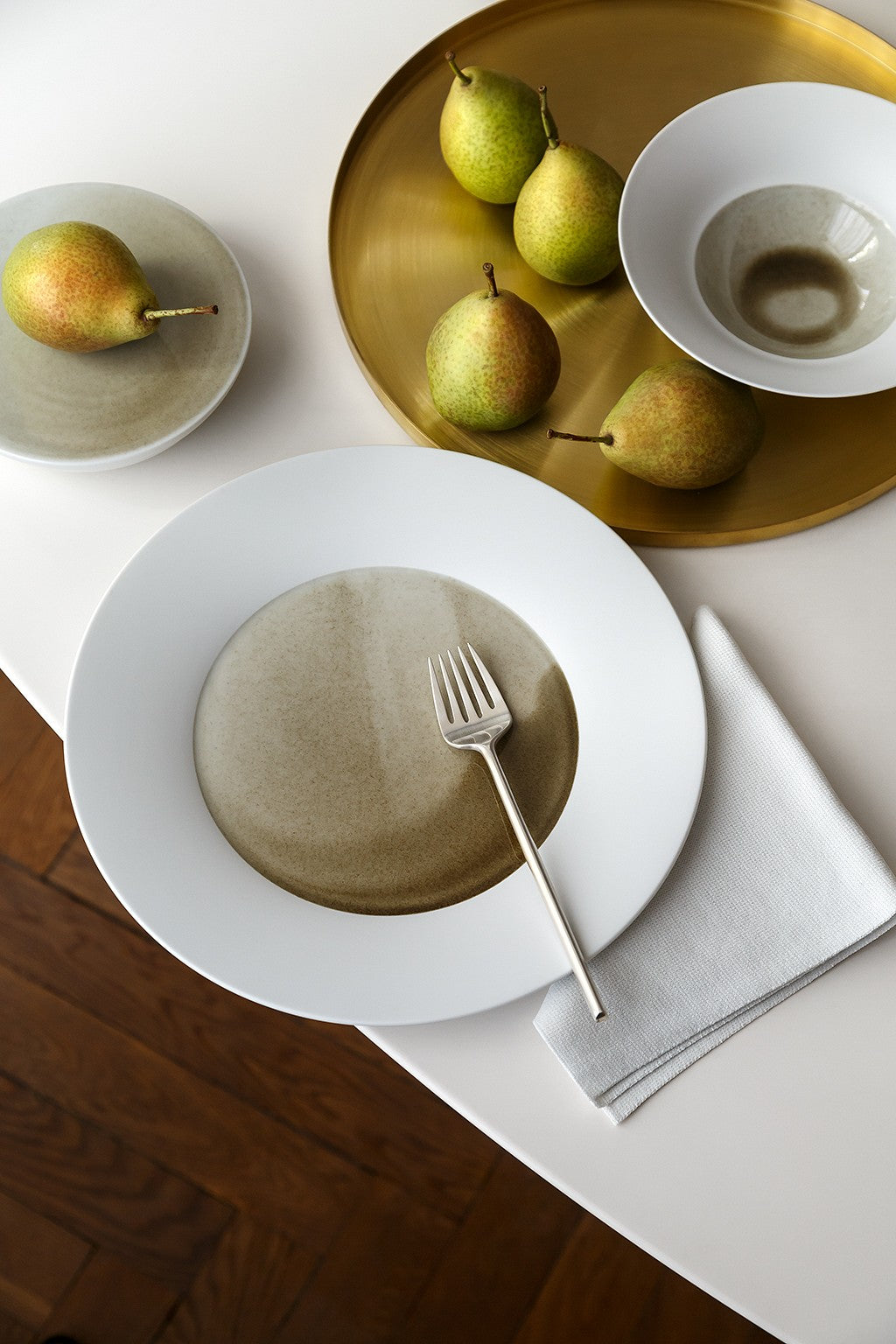 Ein ordentlich gedeckter Esstisch mit einem weißen Teller mit einer Gabel auf einer grauen Serviette, dazu ein Messingtablett mit Birnen und ein weiterer weißer Teller mit einer Birne und einer Teetasse.