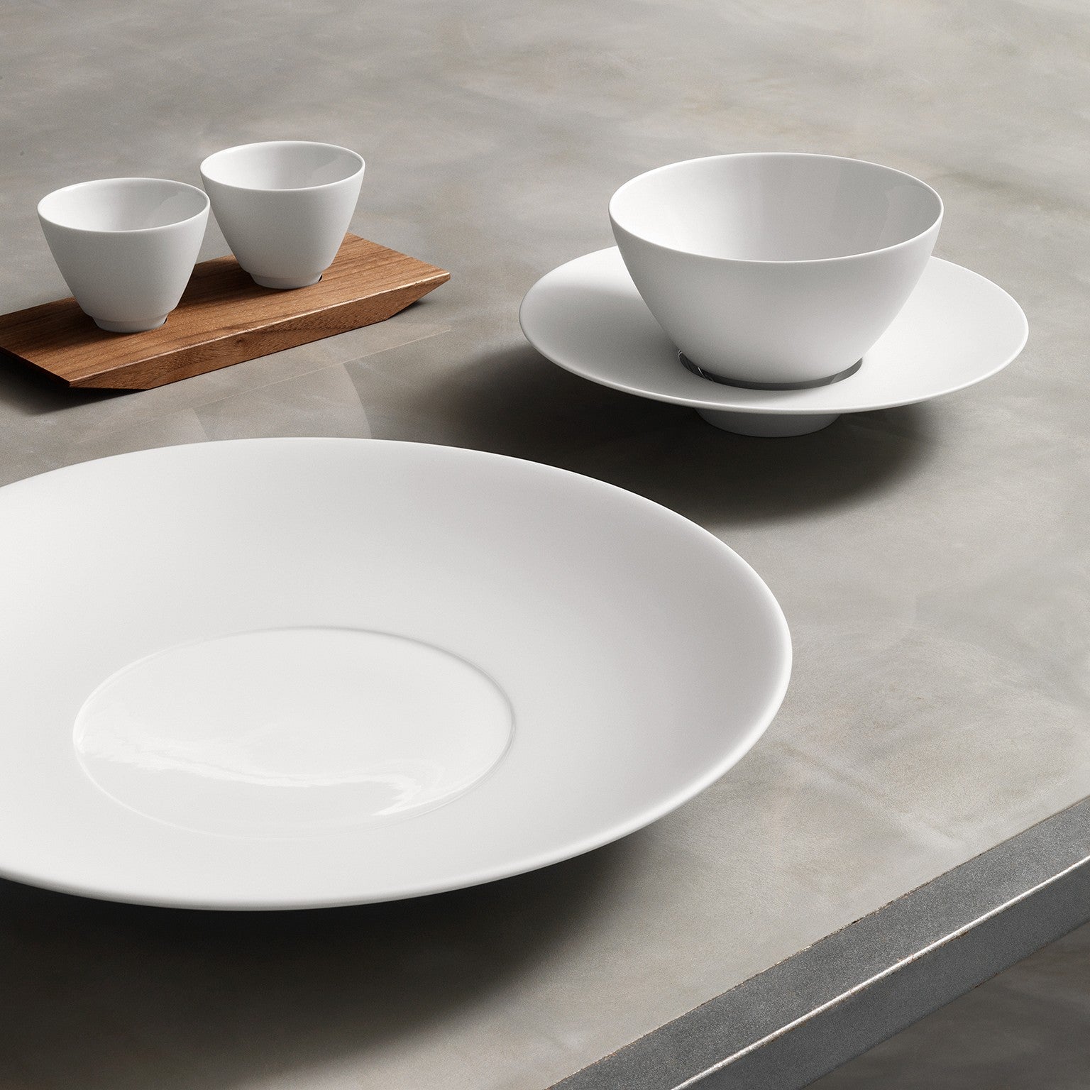 Eine Auswahl an schlichtem weißem Keramikgeschirr, darunter ein großer Teller, zwei Schüsseln unterschiedlicher Größe und ein Paar Tassen, ordentlich angeordnet auf einer glatten grauen Oberfläche mit einem Holzbrett unter den Tassen.