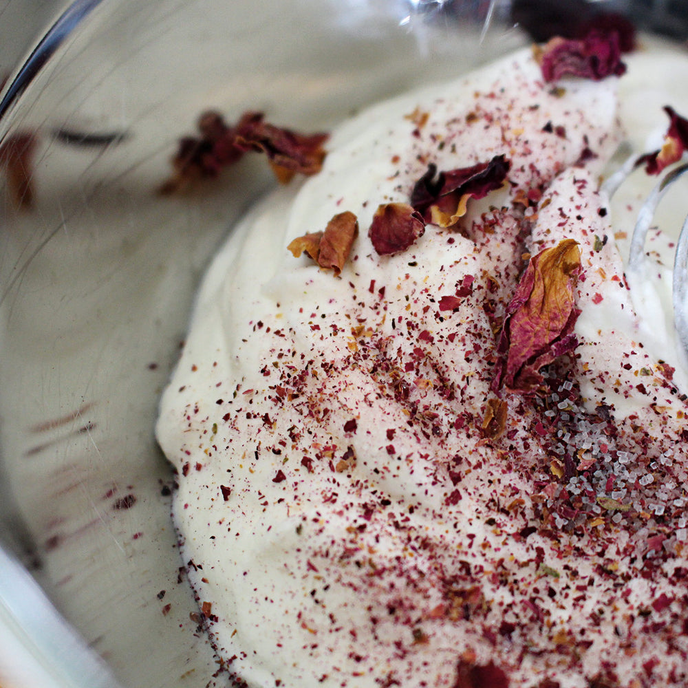 Eine Nahaufnahme von cremigem Joghurt in einer Schüssel, dekoriert mit getrockneten Rosenblütenblättern und einer Prise gemahlener Gewürze. Ein Hauch von Kings Sylter Rosenzucker von der Sylter Manufaktur Johannes King wertet das Gericht mit seinem zarten Geschmack auf. Auf der rechten Seite ist teilweise ein Löffel zu sehen, der neben der Schüssel liegt.