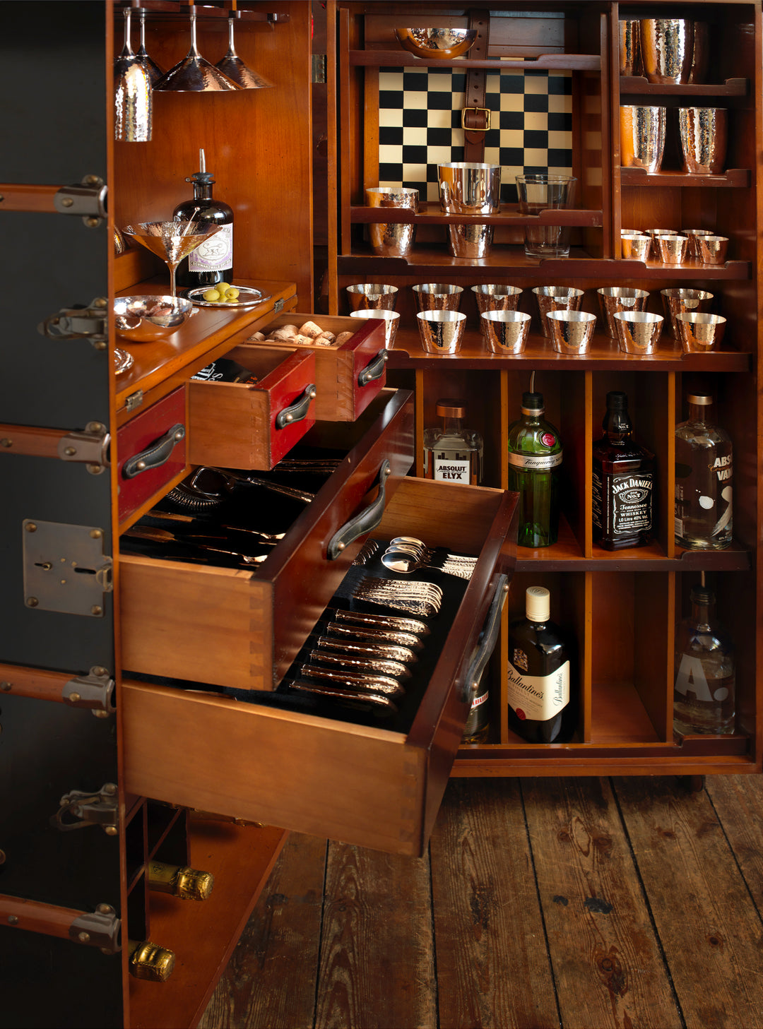 Gezeigt wird ein Holzschrank mit offenen Schubladen, die Besteck enthalten. Die Innenregale enthalten verschiedene Gläser und Alkoholflaschen, darunter Gin, Rum und Whisky. Bekannt als Robbe & Berking Barschrank im Kofferdesign schwarz – mit Ausstattung von Robbe & Berking, verfügt er über Fächer mit Bar-Zubehör wie Cocktail-Shakern und ein Schachbrettmuster an der Rückwand.