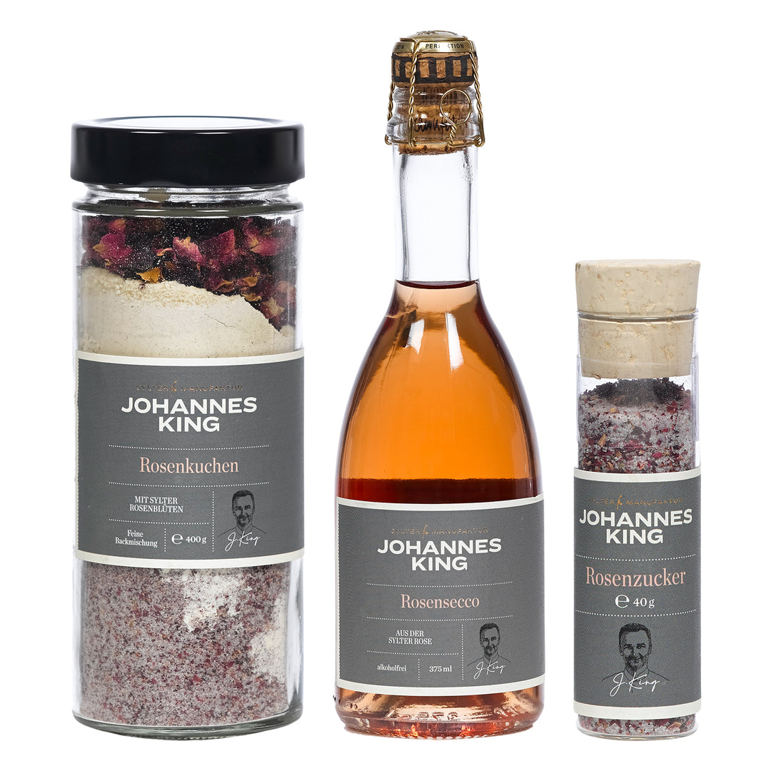 Ein Produktbild zeigt drei exquisite Spezialitäten der Sylter Manufaktur Johannes King: ein Glas Rosenkuchenmischung, eine Flasche Rosensecco und einen kleinen Behälter Rosenzucker. Die geschmackvolle graue Verpackung zeigt Text und Johannes Kings Unterschrift und zeigt die Eleganz der „Süßen rosigen Verführung von der Insel“.