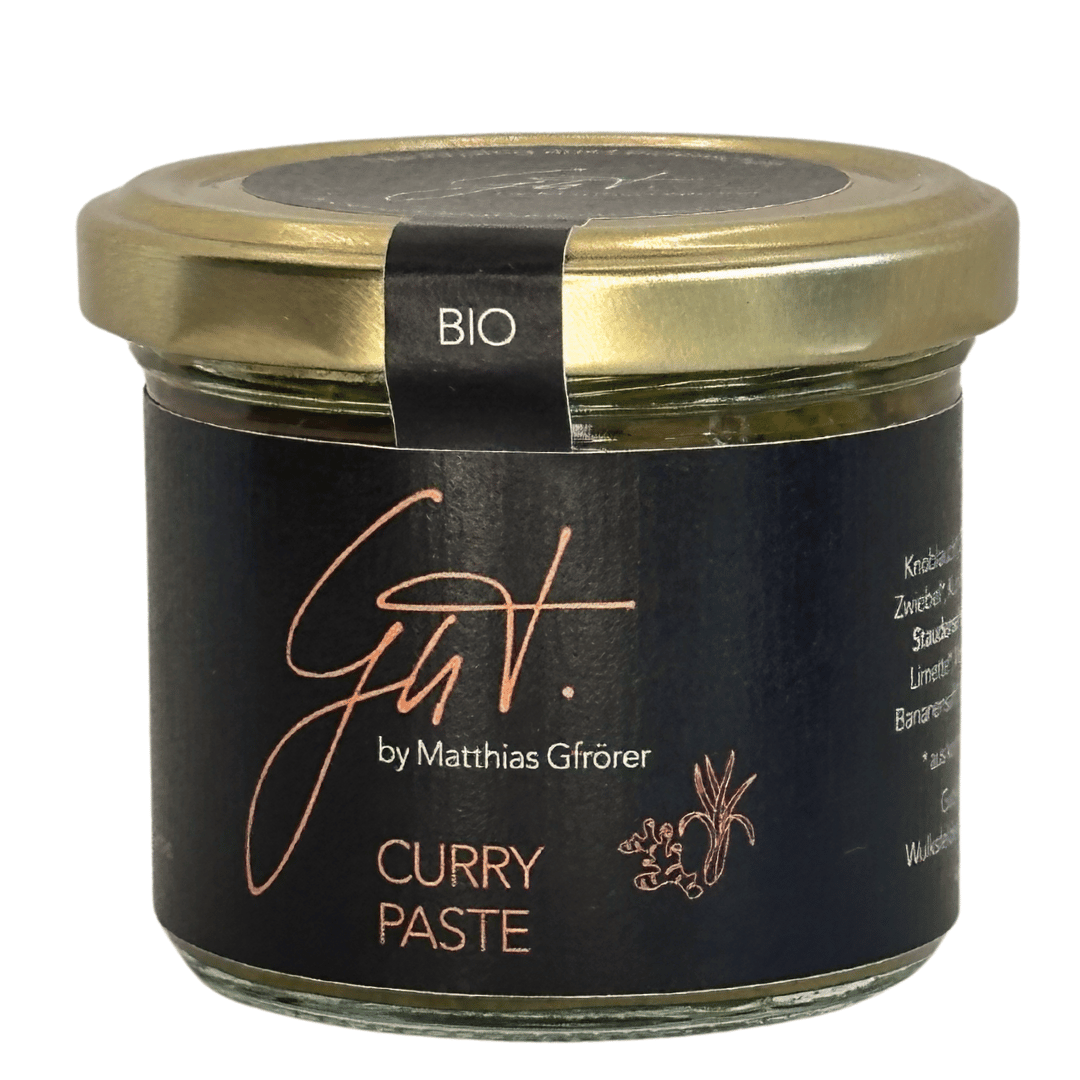 Ein Glasgefäß mit „Matthias Gfrörers Bio Curry-Paste“ mit schwarzem Etikett und goldenem Deckel, isoliert auf weißem Hintergrund. Der Text auf dem Glas enthält die Zutaten und stammt von Matthias Gfrörer.