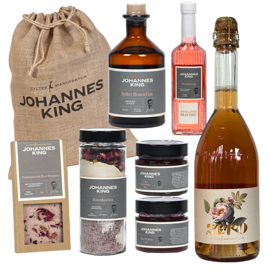 Die Kollektion „Großer rosiger Nikolaus“ der Sylter Manufaktur Johannes King umfasst einen Leinenbeutel, Sylter Rosen Gin, einen rosa Likör, Rosensekt in einer großen Flasche mit Blumenetikett, verschiedene Gläser mit Marmelade oder Aufstrichen und ein Paket mit Brot oder Keksen – alles einheitlich beschriftet.